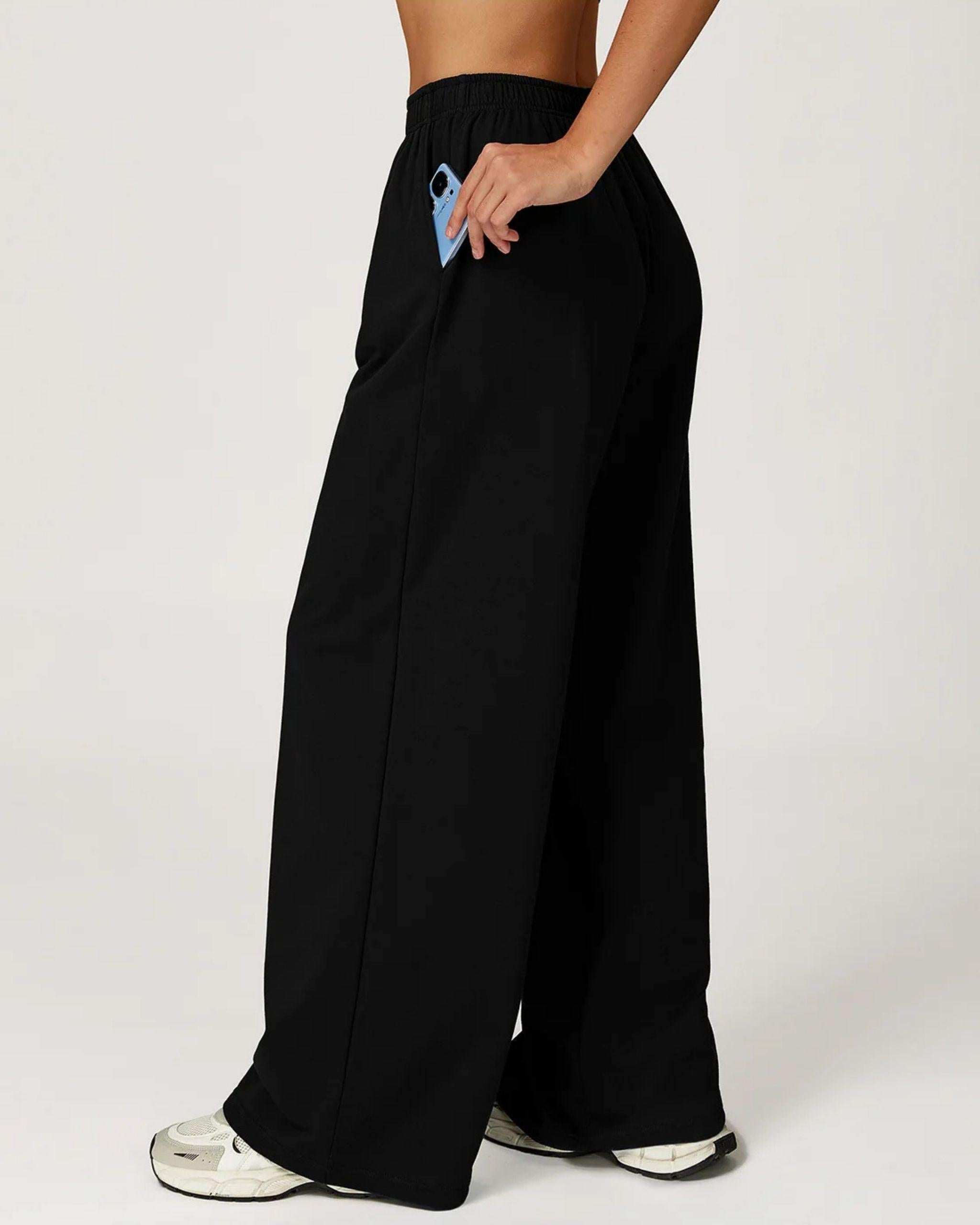 Seitenansicht der schwarzen Wide Leg Sporthose, Hand steckt ein blaues Handy in die funktionale Seitentasche, elastischer High-Waist Bund ist sichtbar - Ryze.